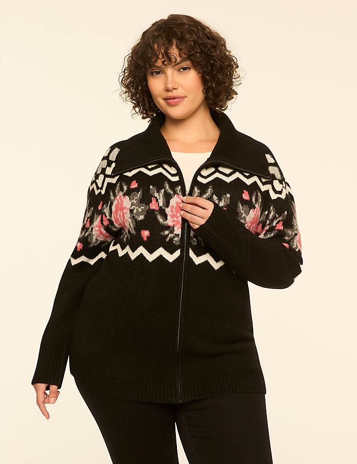 A woman with curly brown hair wearing a black floral zip-up cardigan and black pants stands against a plain off-white background.