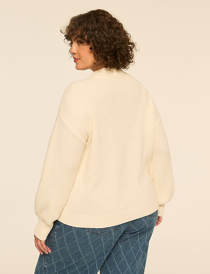 A woman with curly brown hair wearing a cream-colored ribbed sweater and blue jeans stands against a plain, light-beige background.