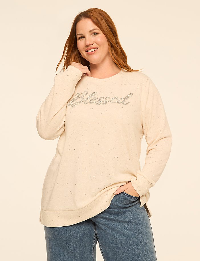 A smiling woman wearing a cream-colored "blessed" sweater and blue jeans stands against a plain, light-colored background.