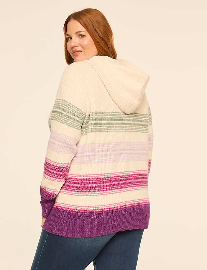 A woman with red hair wearing a hooded, multi-colored striped sweater and blue jeans stands against a plain, light-colored background.