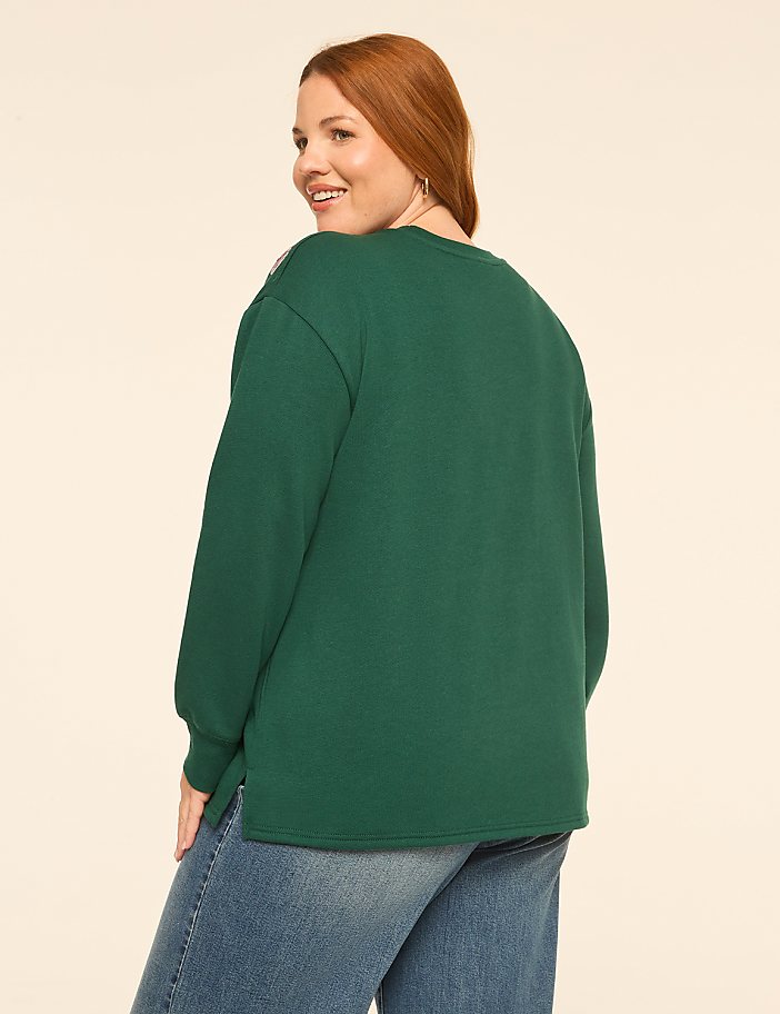 A woman with red hair wearing a green sweatshirt and blue jeans stands against a plain, light-colored background.