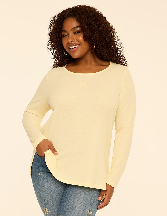 A smiling woman with curly hair wearing a pale yellow long-sleeved shirt and blue jeans stands against a plain, light-colored background.