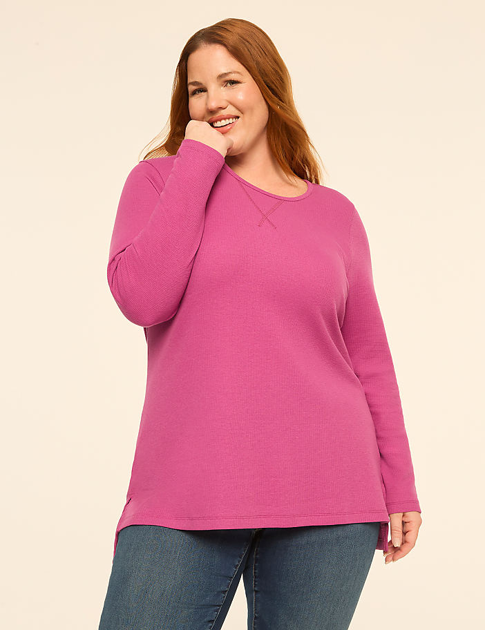 A smiling woman with red hair wearing a bright pink long-sleeved shirt and blue jeans stands against a plain, light-colored background.