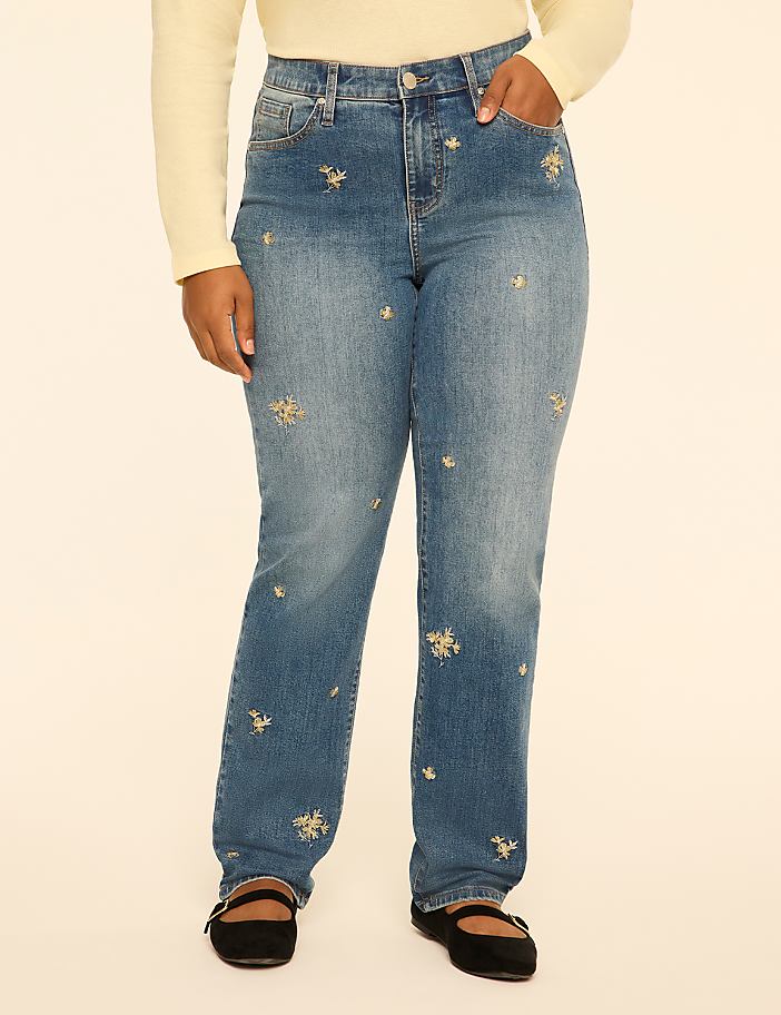 A person wearing a cream-colored top, blue jeans with floral embroidery, and black flats stands against a plain white background.