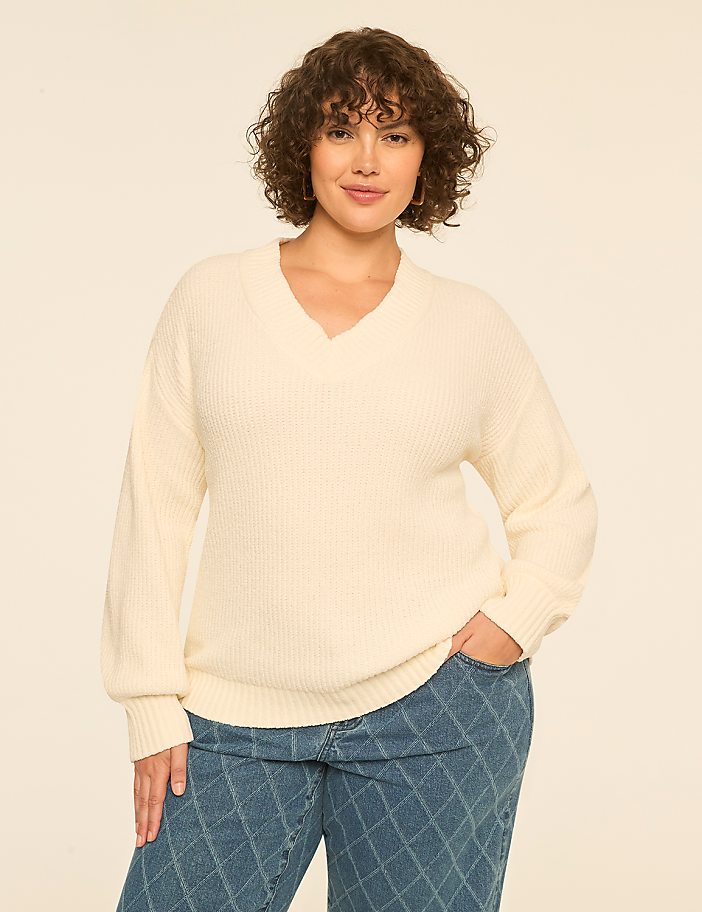 A woman with curly brown hair wearing a cream v-neck sweater and patterned blue jeans stands against a plain off-white background.