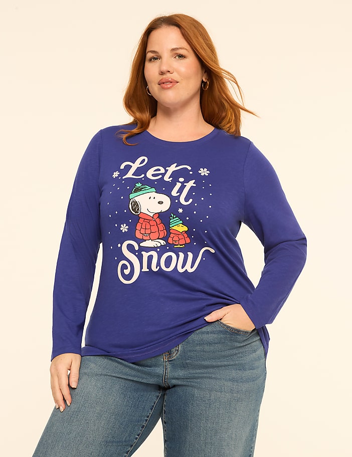 A woman with red hair wearing a blue "Let it Snow" Snoopy shirt and blue jeans stands against a plain off-white background.