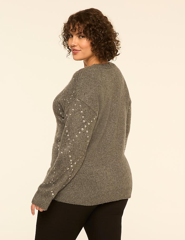 A woman with curly hair wearing a grey embellished sweater and black pants stands against a plain white background.