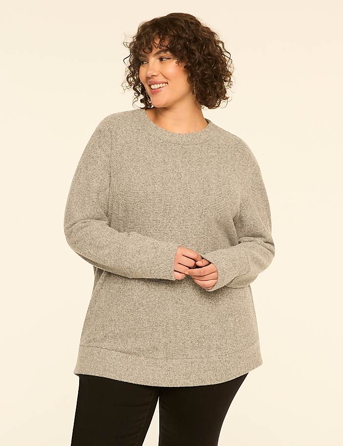 A smiling woman with curly brown hair wearing a grey sweater and black pants stands against a plain off-white background.