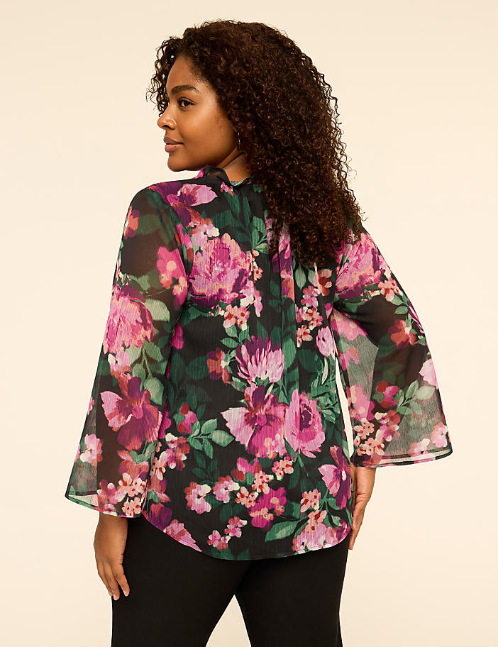 A woman with curly hair wearing a floral print blouse and black pants stands against a plain, light-colored background.