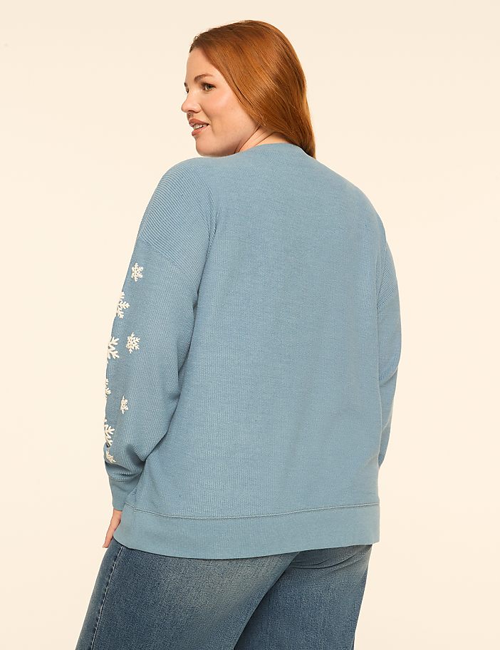 A woman with red hair wearing a blue snowflake-patterned sweater and jeans stands against a plain white background.