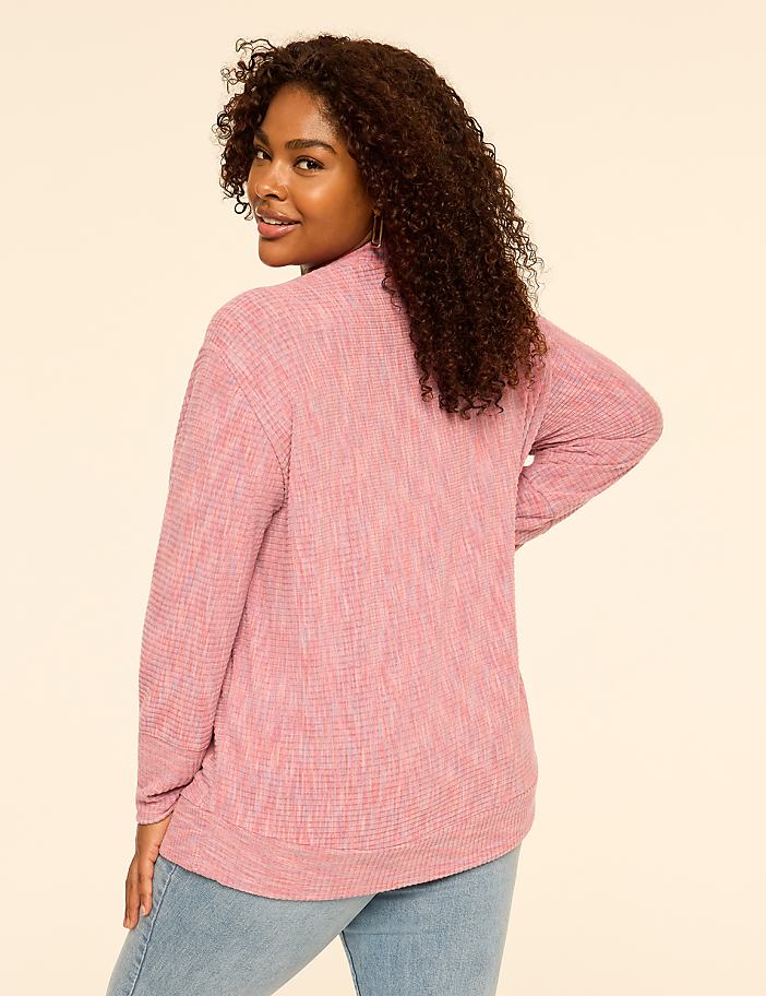 A woman with curly brown hair wearing a pink textured top and blue jeans stands against a plain, light-colored background.