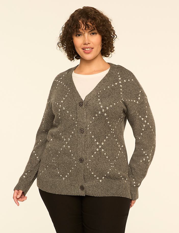 A woman with curly hair wearing a grey embellished cardigan and black pants stands against a plain off-white background.