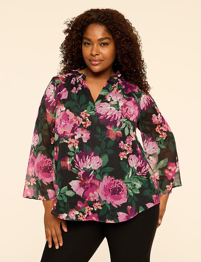 A woman with curly hair wearing a floral print blouse and black pants stands against a plain, light-colored background.