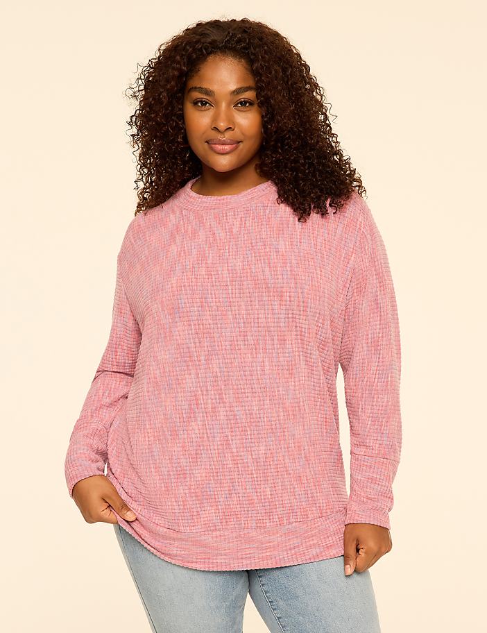 A woman with curly hair wearing a pink textured long-sleeve top and light blue jeans stands against a plain, light-colored background.