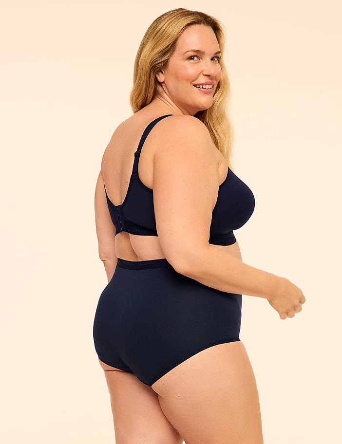 A smiling blonde woman wearing a navy blue bra and high-waisted briefs stands against a plain, light beige background.