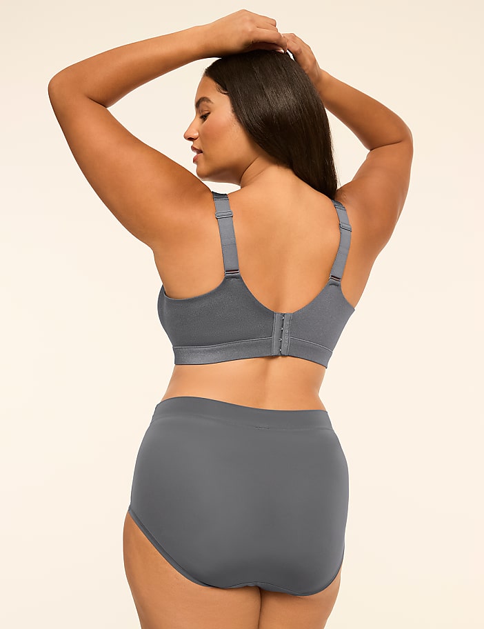 A woman wearing a grey bra and matching high-waisted briefs stands with her arms raised against a plain, light-beige background.