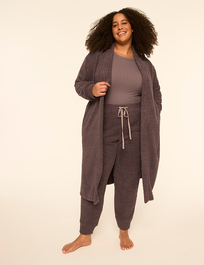 A smiling woman with curly hair wears a mauve-colored lounge set and long robe against a plain, light-colored background.