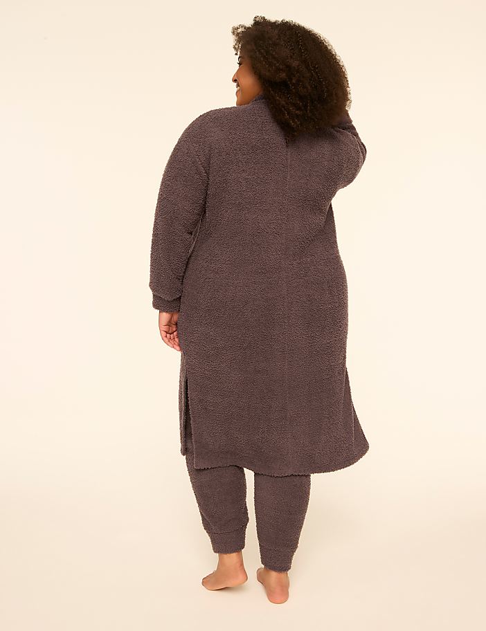 A woman with curly hair wearing a dark purple fuzzy robe and matching pants stands against a plain, light-colored background.