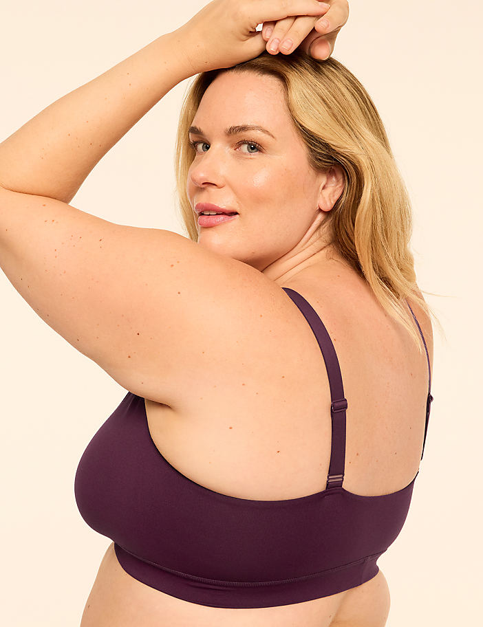 A blonde woman in a purple bra poses with her arms raised against a plain, light-colored background.