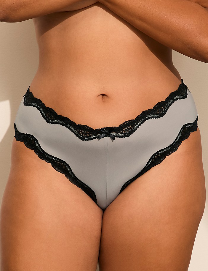 A person wears grey panties with black lace trim against a plain, neutral-colored wall.