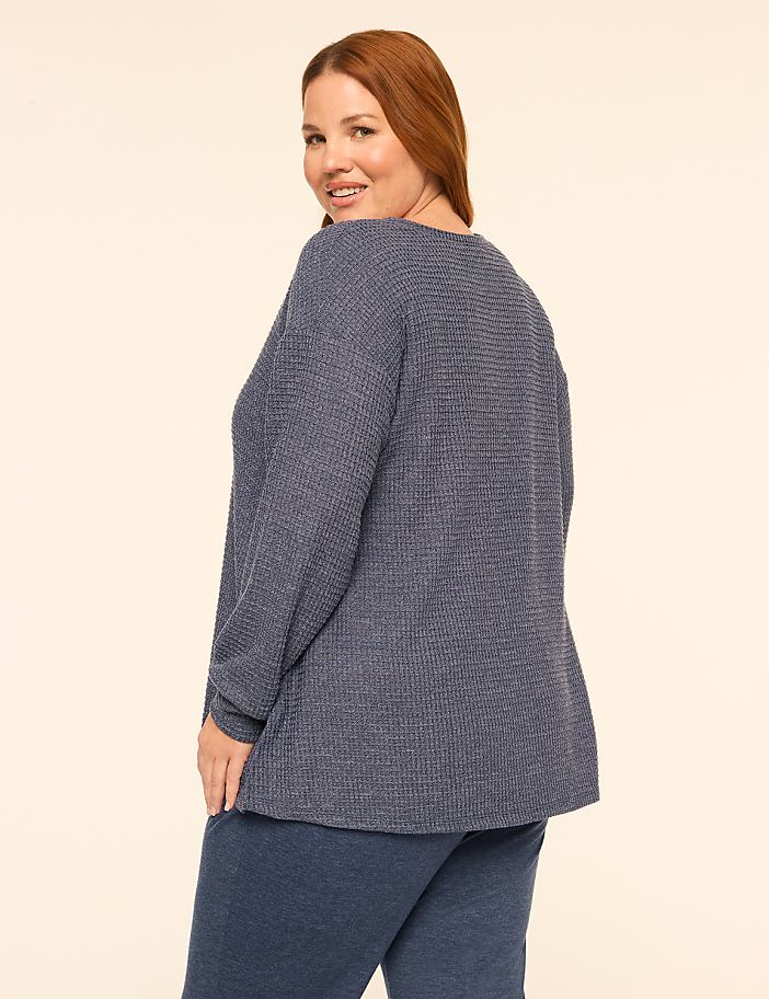 A woman with red hair wearing a blue textured long-sleeve top and matching pants stands against a plain, light-colored background.