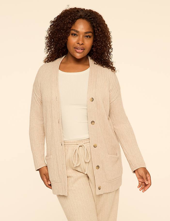 A woman with curly hair wearing a beige cardigan and matching pants stands against a plain, light-colored background.