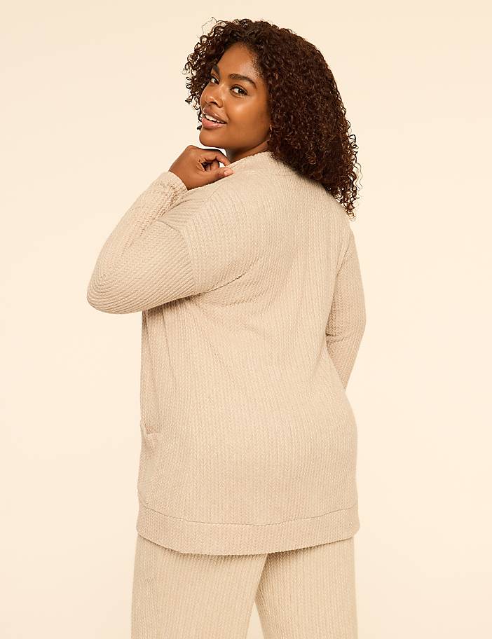 A woman with curly hair wearing a beige ribbed lounge set poses against a plain, light-colored background.
