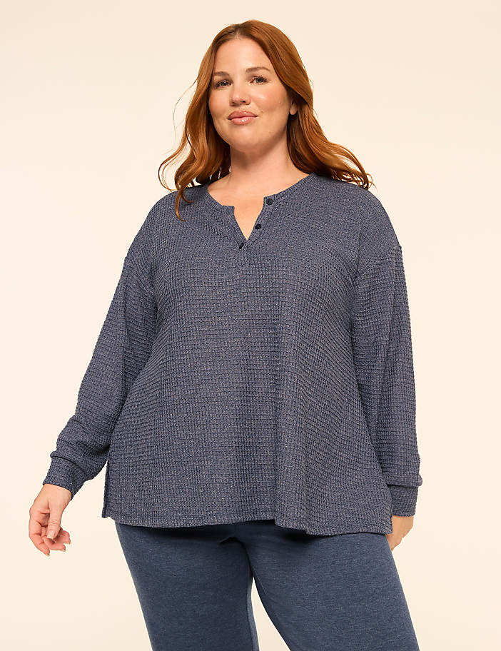 A woman with red hair wearing a blue textured long-sleeve top and matching pants stands against a plain, light-colored background.