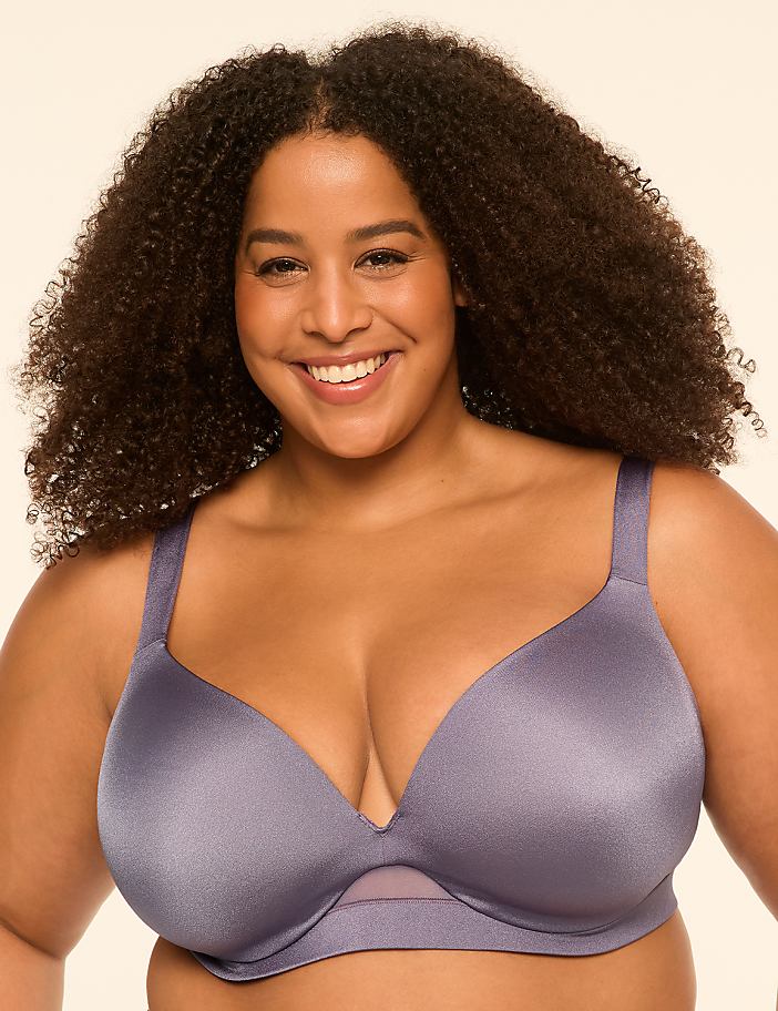 A smiling woman with curly brown hair wearing a purple bra stands against a plain, light-colored background.