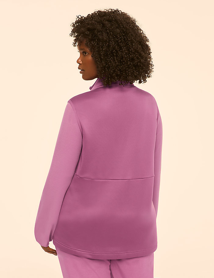 A woman with dark curly hair stands facing away from the camera, wearing a mauve jacket and pants against a plain, light-colored background.