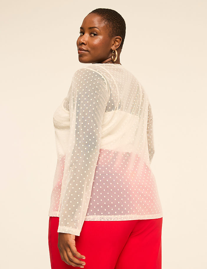 A woman wearing a sheer white polka-dot top and red pants stands against a plain, light-colored background.
