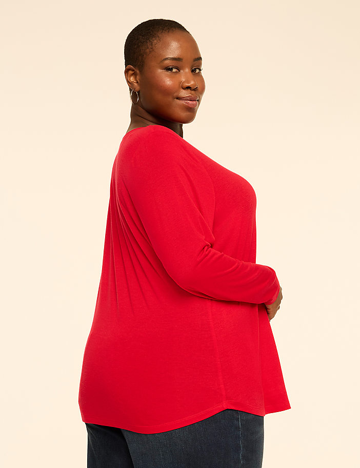 A woman wearing a red long-sleeved top and dark blue jeans poses against a plain, light-colored background.