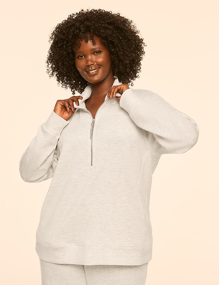 A smiling woman with dark curly hair poses in a light-colored quarter-zip sweatshirt against a plain, light background.