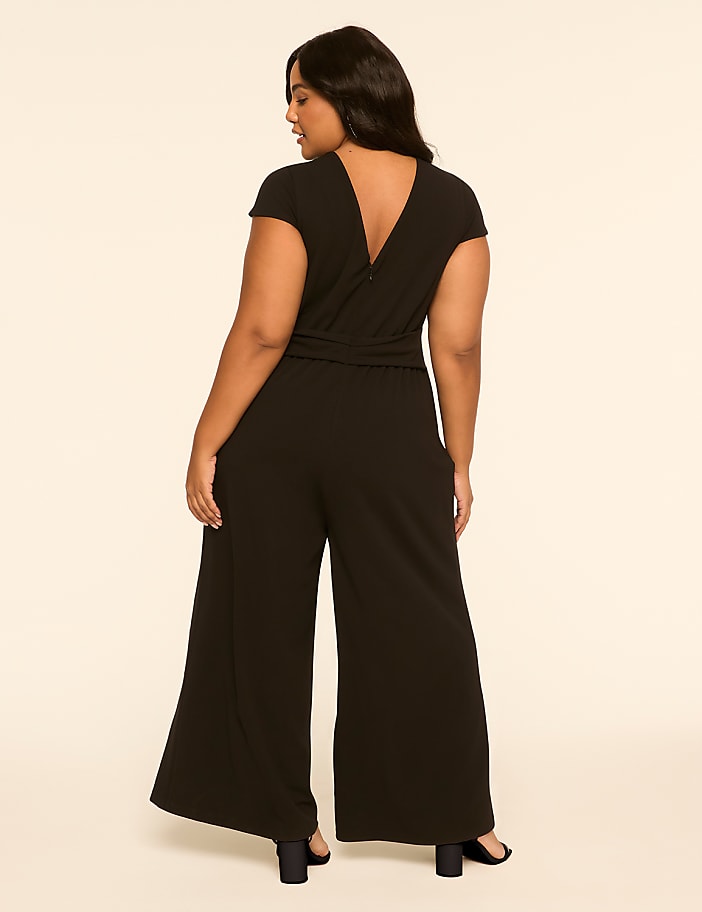A woman in a black jumpsuit stands against a plain, light-colored background.