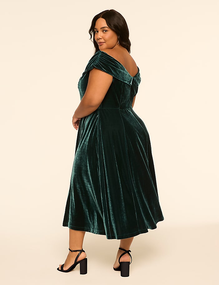 A woman in a green velvet dress stands against a plain, light-colored background.