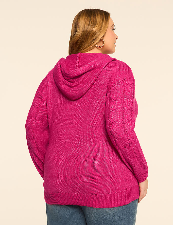 A woman in a pink hooded sweater and jeans stands against a plain, light-colored background.