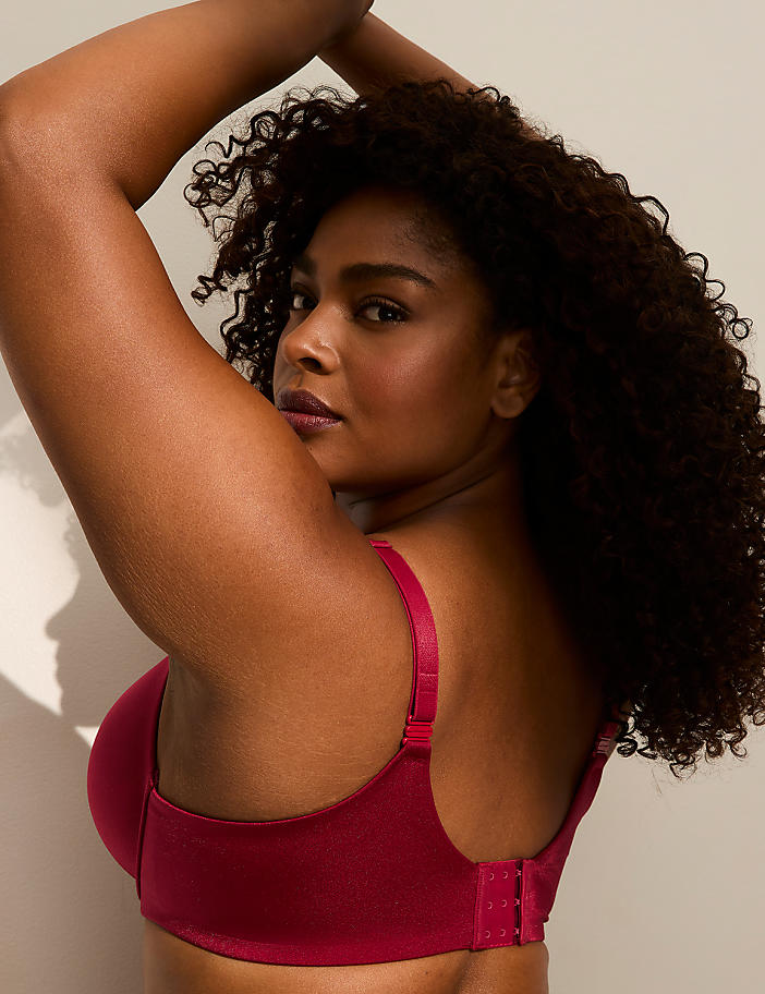 A woman with curly dark hair wearing a red bra poses with one arm raised against a plain, light-colored wall.