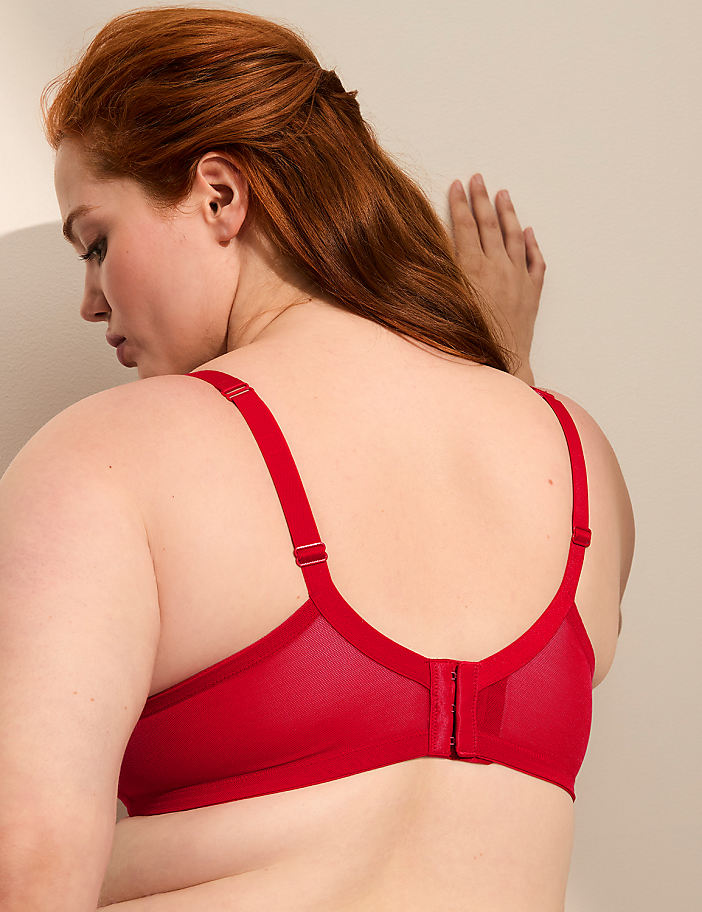 A woman with red hair wearing a red bra stands against a plain, light-colored wall.