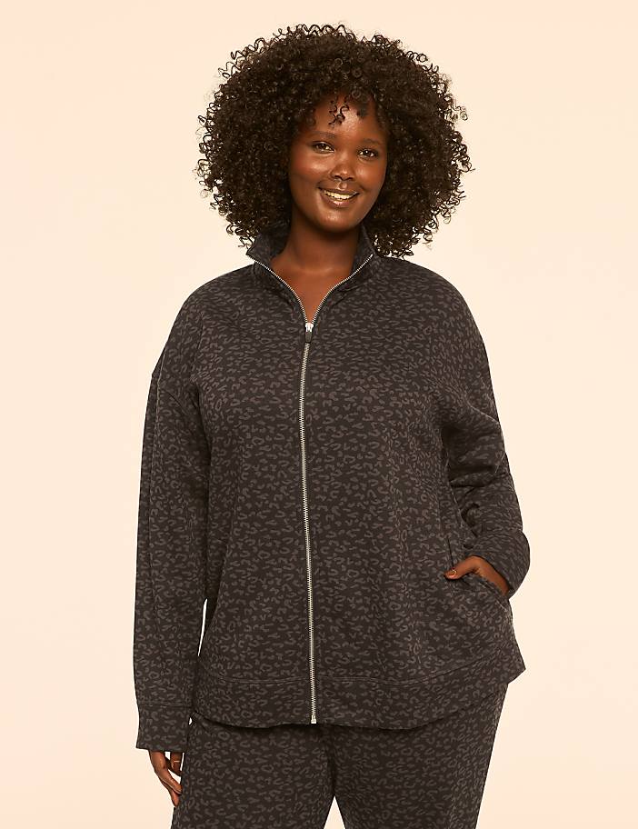 A smiling woman with curly dark hair stands in the foreground wearing a leopard-print zip-up jacket and matching pants against a plain, light-colored background.