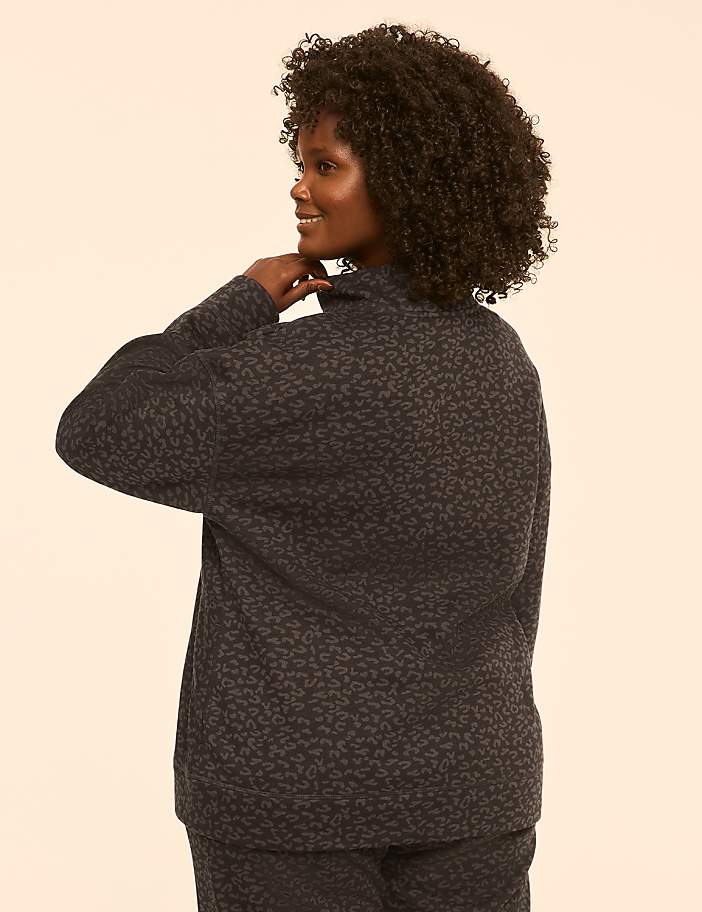 A woman with curly dark hair, wearing a patterned dark top and pants, is shown from the back with her hand on her neck against a plain, light-colored background.
