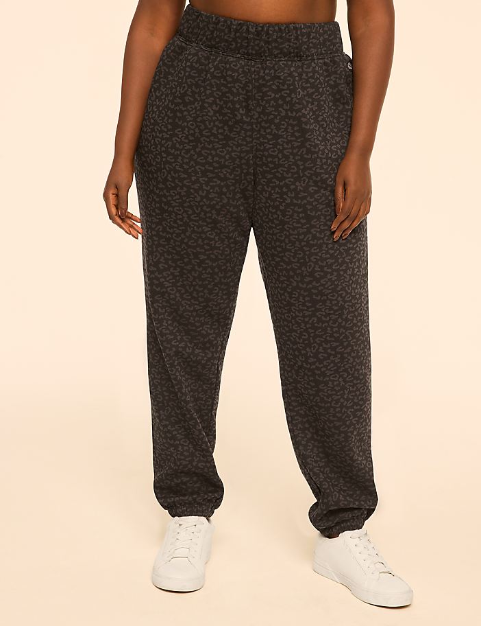 A person wearing leopard-print sweatpants and white sneakers stands against a plain, peachy-toned background.