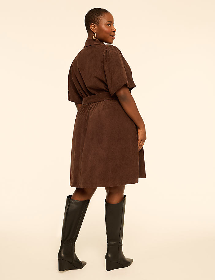 A woman in a brown dress stands against a plain, light-colored background.