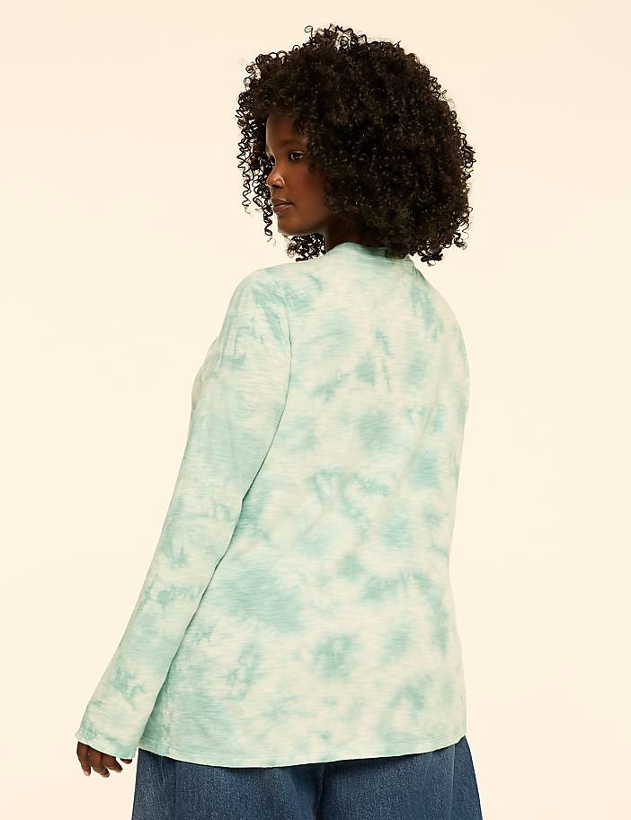 A woman with curly hair poses in a teal and mint green ombre shirt against a plain cream background.