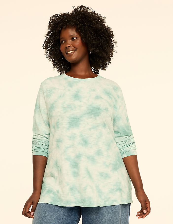 A woman with curly hair smiles at the camera, wearing a long-sleeved, ombre teal shirt and jeans against a plain, light-colored background.