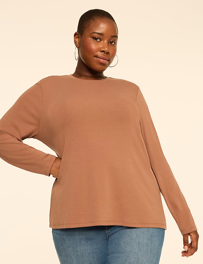 A woman wearing a brown long-sleeved shirt and blue jeans stands against a plain, light-colored background.