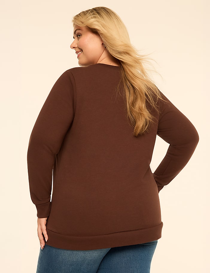 A blonde woman wearing a brown long-sleeved shirt and blue jeans stands against a plain, light-colored background.