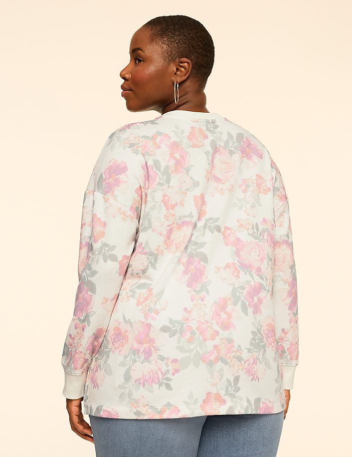 A woman wearing a floral print long-sleeve shirt and blue jeans stands against a plain, light-colored background.