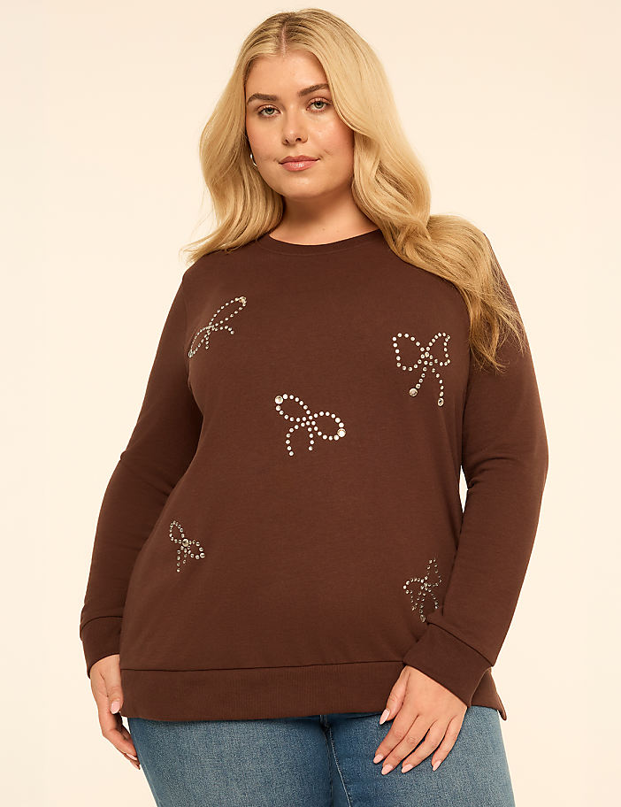 A blonde woman wearing a brown sweatshirt with rhinestone bows and blue jeans stands against a plain, light-colored background.