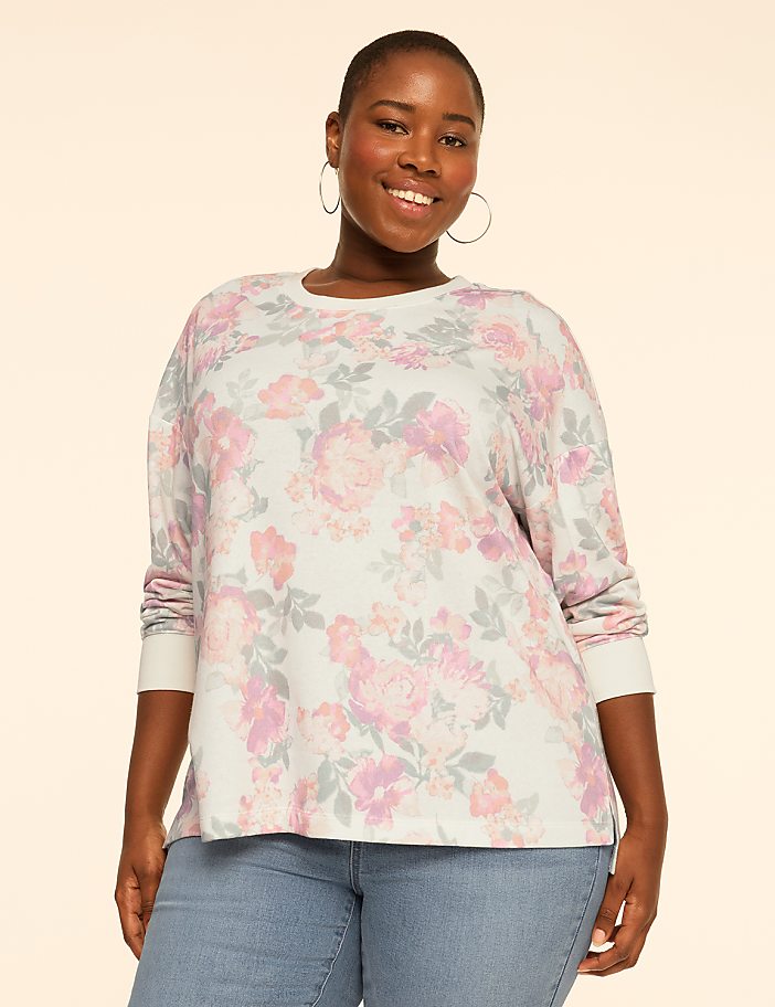 A smiling woman wearing a floral print top and blue jeans stands against a plain, light-colored background.