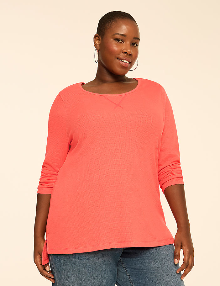 A woman wearing a coral-colored long-sleeved shirt and blue jeans stands against a plain, light beige background.